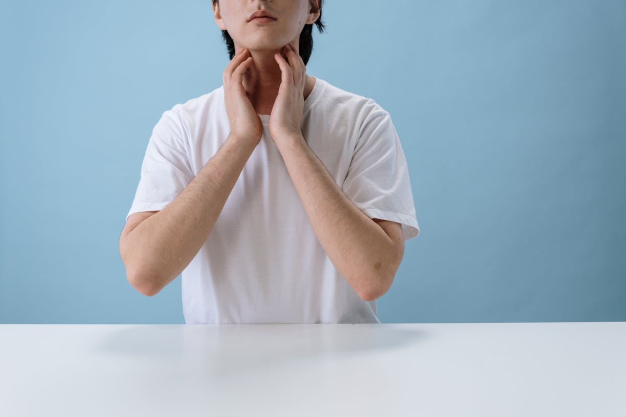Home Close-up of an individual in a white crew neck t-shirt placing hands near neck, isolated on a blue backdrop.