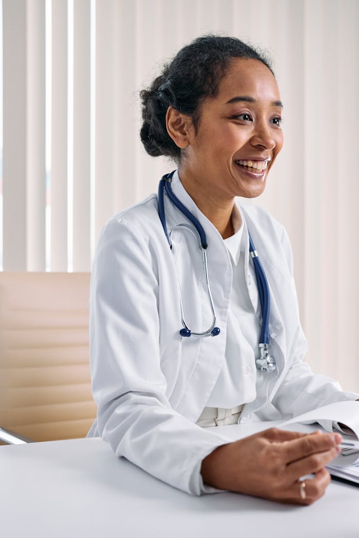 About Black female doctor smiling at desk with stethoscope, in a bright medical office.